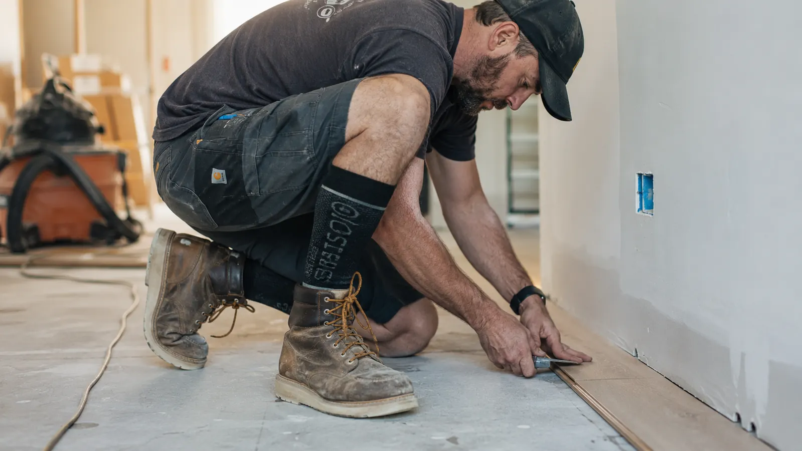 Flooring installer crouching on a concrete subfloor, compression socks visible above work boots, mid-project
